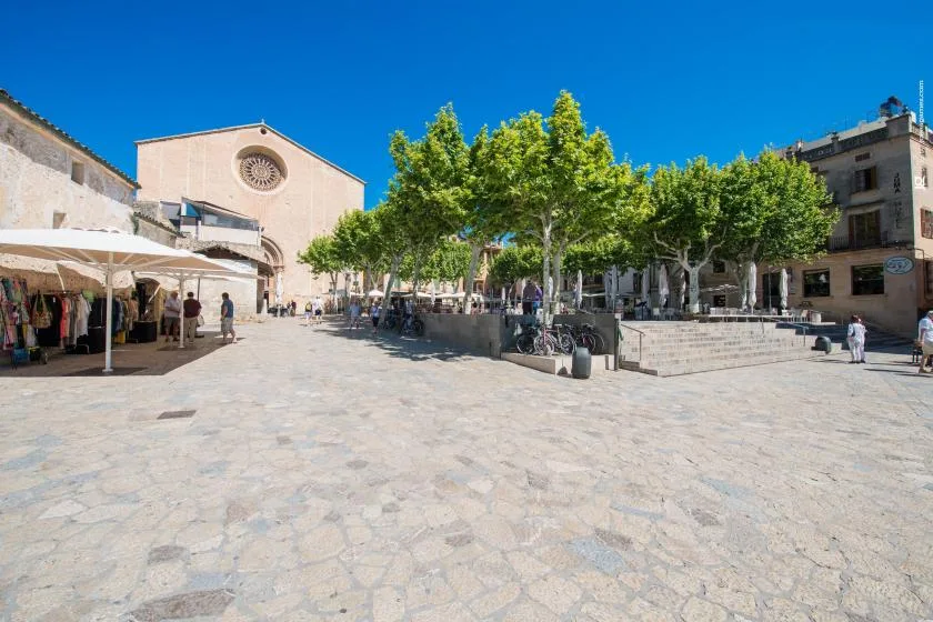 Una plaza pública soleada con árboles frondosos, puestos de mercado y edificios alrededor.
