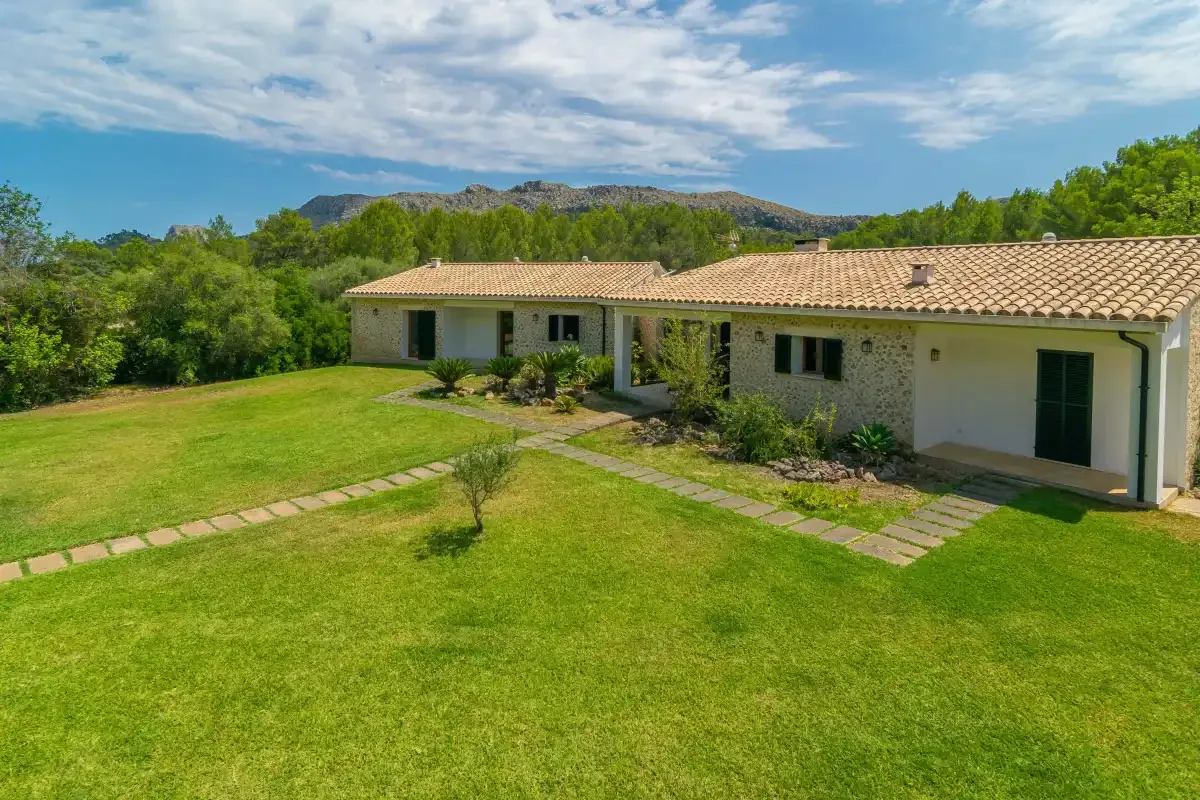 Una casa baja junto a un amplio césped verde, con árboles y colinas al fondo.