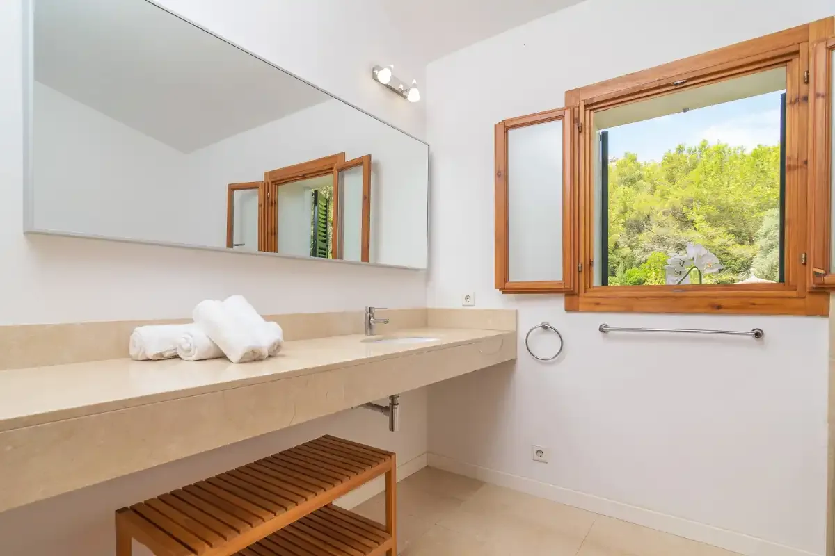 Un lavabo de baño con un espejo grande y una ventana abierta con vistas a los árboles.