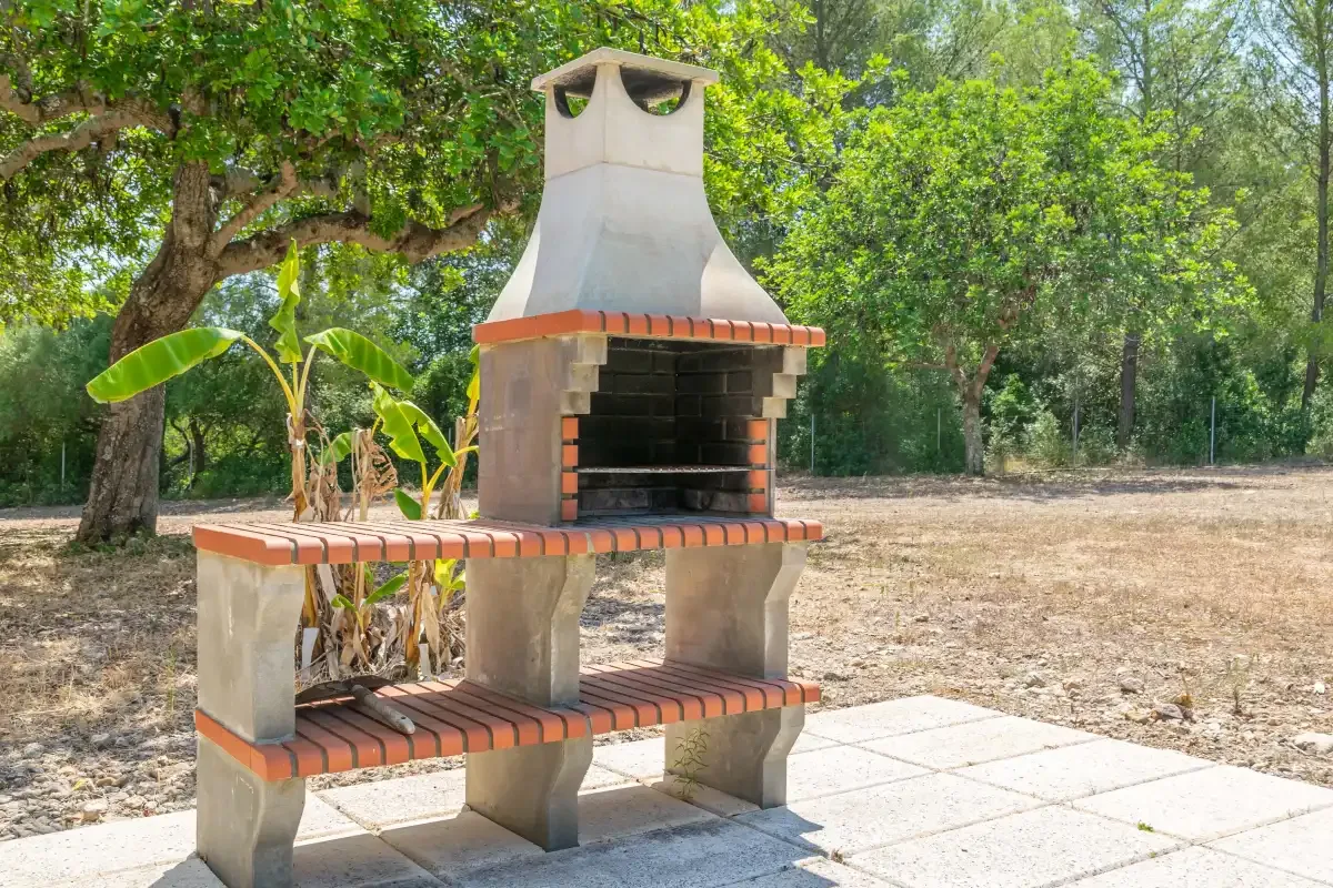 Una parrilla exterior está bajo árboles frondosos, junto a una zona pavimentada.