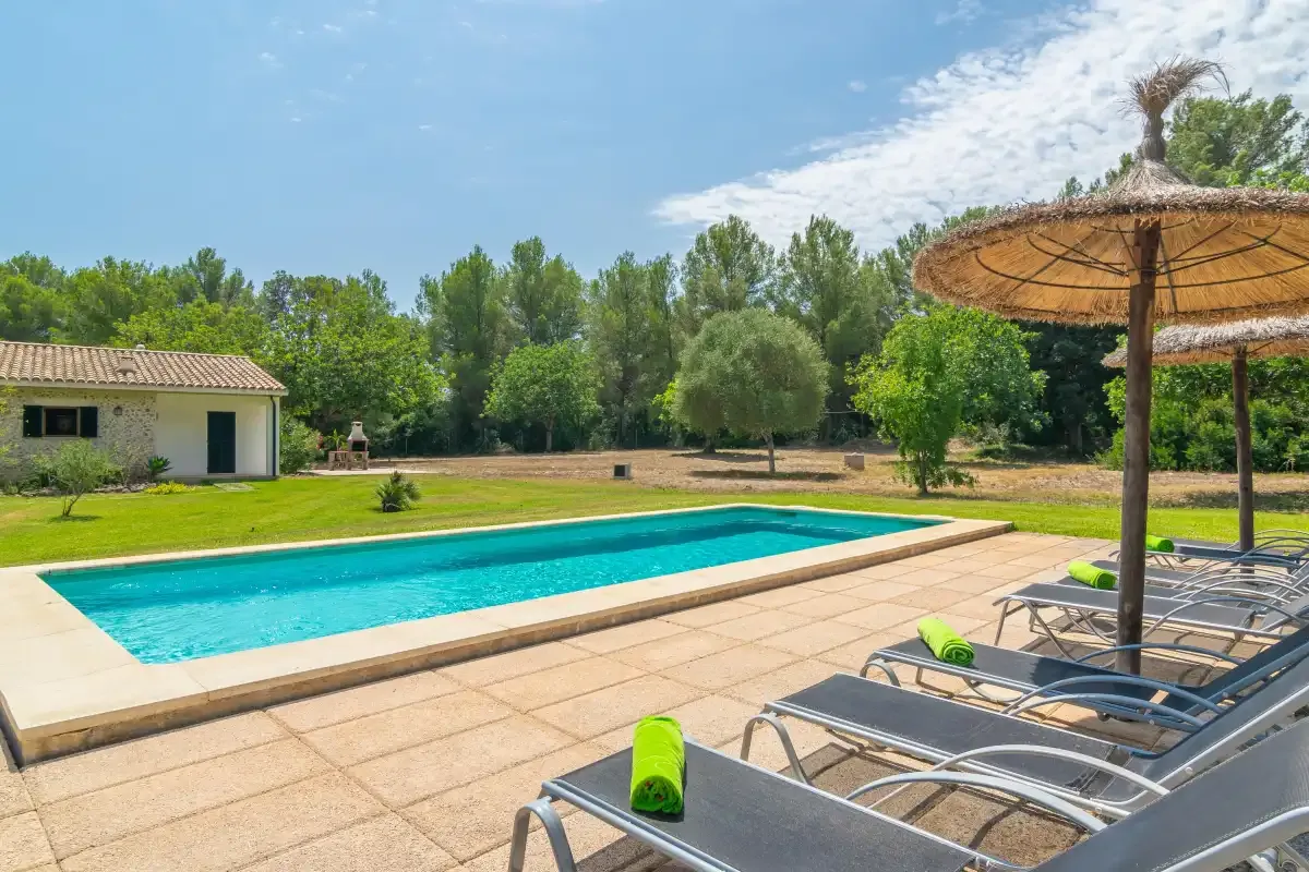 Una piscina junto a un patio pavimentado con tumbonas y una sombrilla de paja.