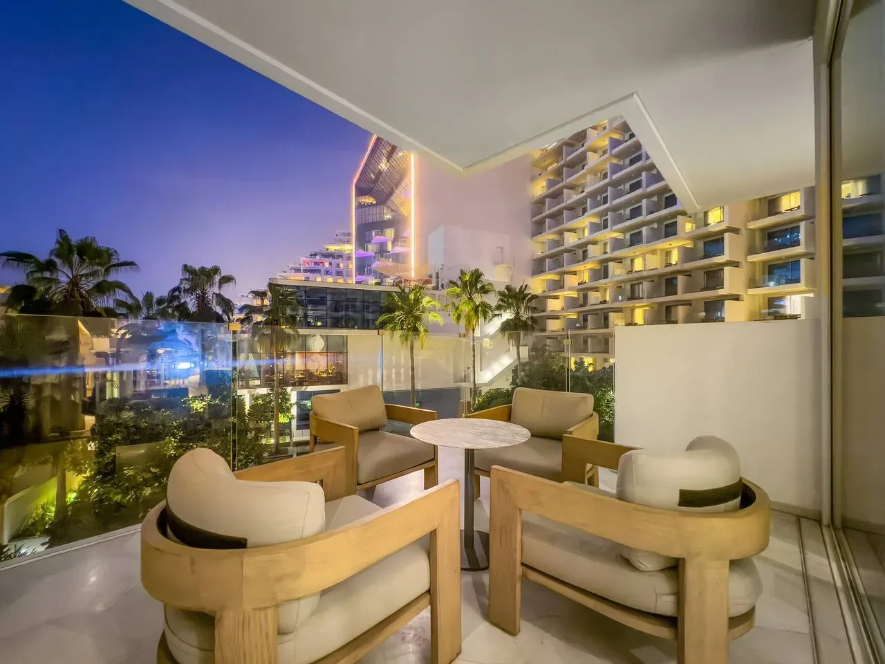 Una zona de estar en el balcón da a palmeras y edificios iluminados al anochecer.