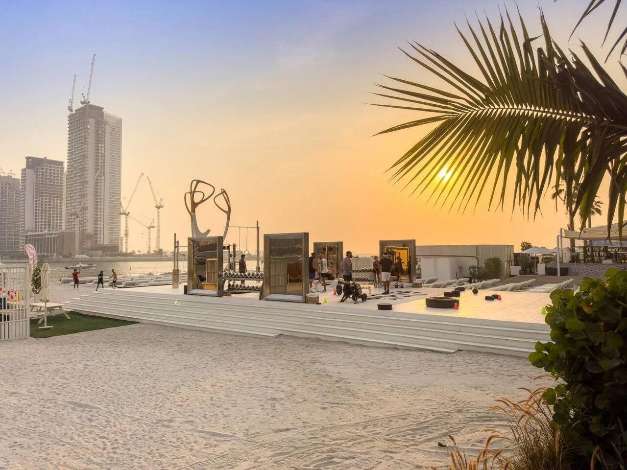Una zona de eventos junto a la playa al atardecer, con exposiciones y edificios altos a lo lejos.