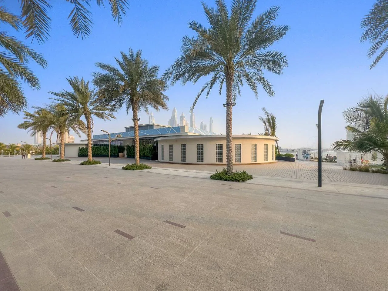 Un edificio moderno bajo se encuentra junto a una amplia plaza pavimentada con palmeras.