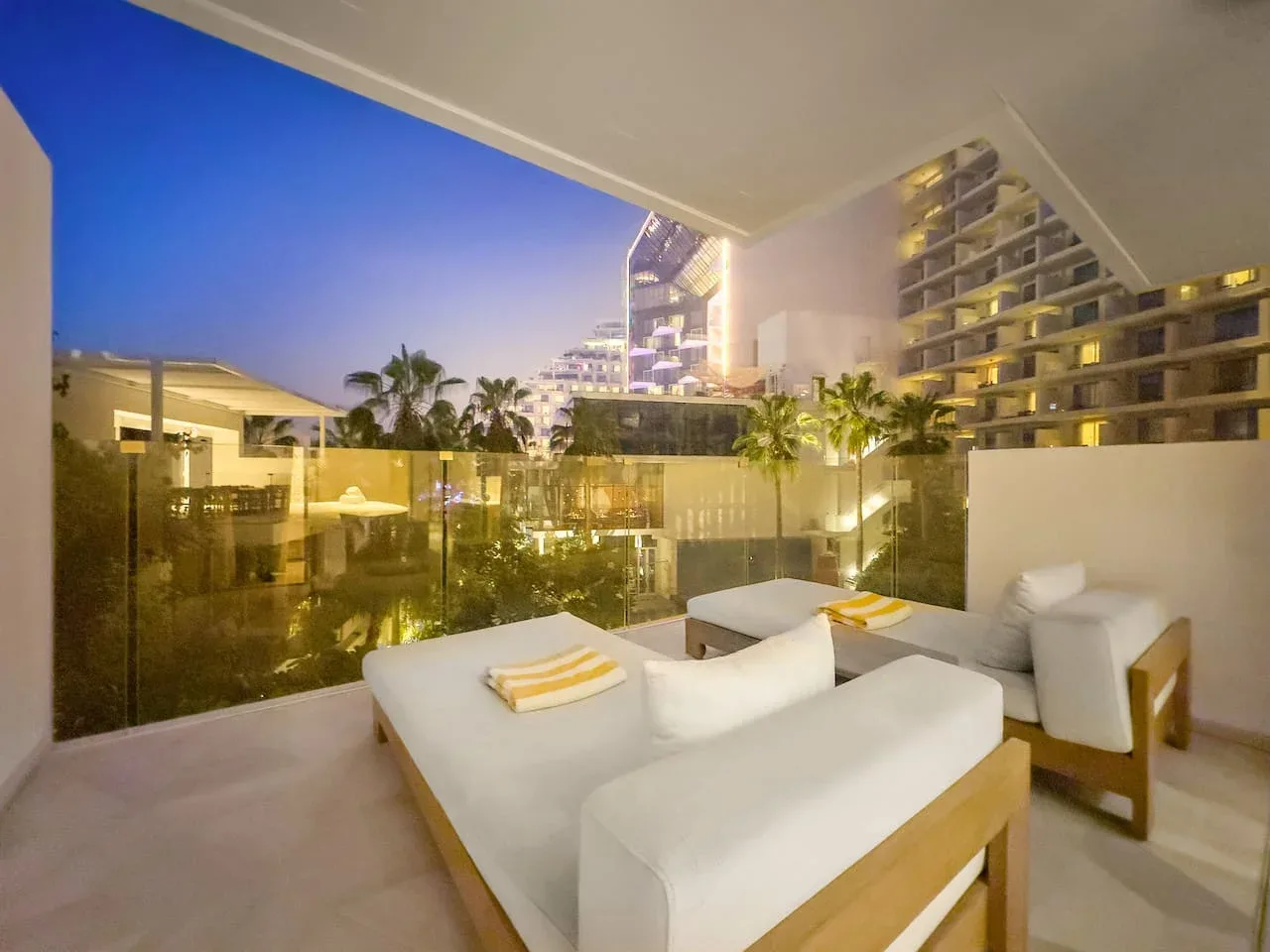 Una terraza con asientos blancos ofrece vistas de edificios iluminados y palmeras al anochecer.