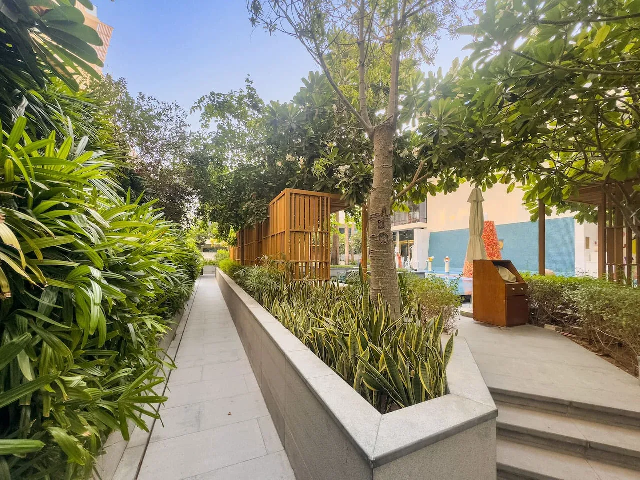 Un patio ajardinado tiene senderos pavimentados, vegetación abundante y una celosía de madera.