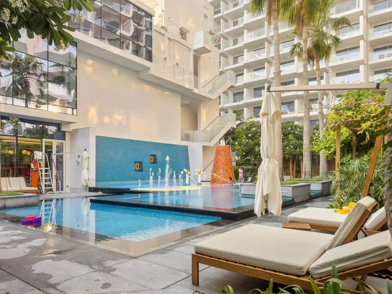 Una piscina al aire libre junto a un edificio moderno, con tumbonas y palmeras.