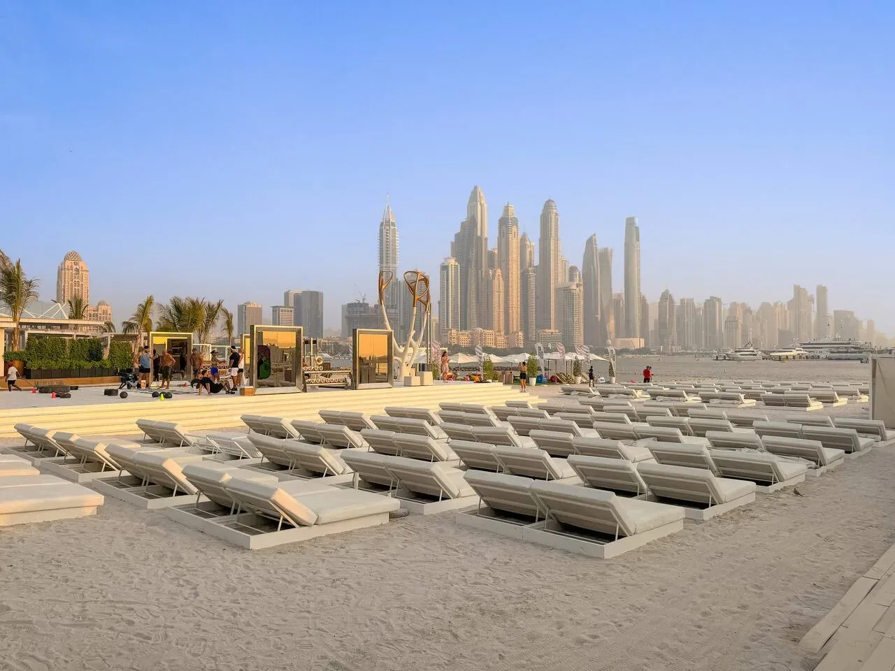 Una playa de arena junto al agua está bordeada de tumbonas, con el perfil urbano a lo lejos.