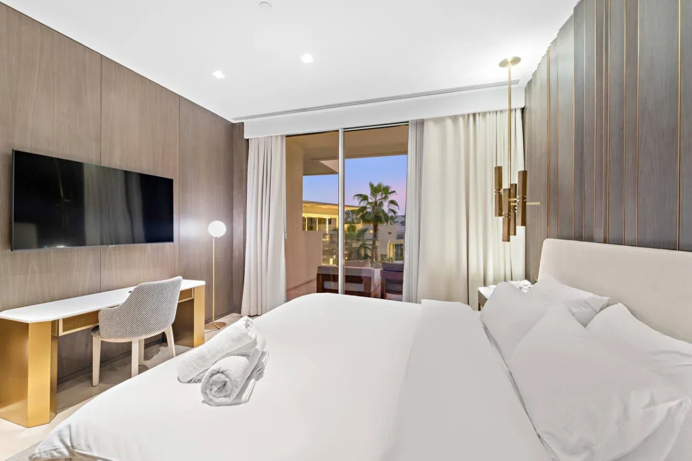 Un dormitorio luminoso con una cama grande, un escritorio y una ventana con vista a palmeras.
