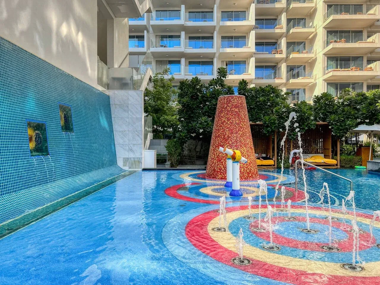 Una colorida zona de piscina al aire libre tiene fuentes de agua y una pared de mosaico.