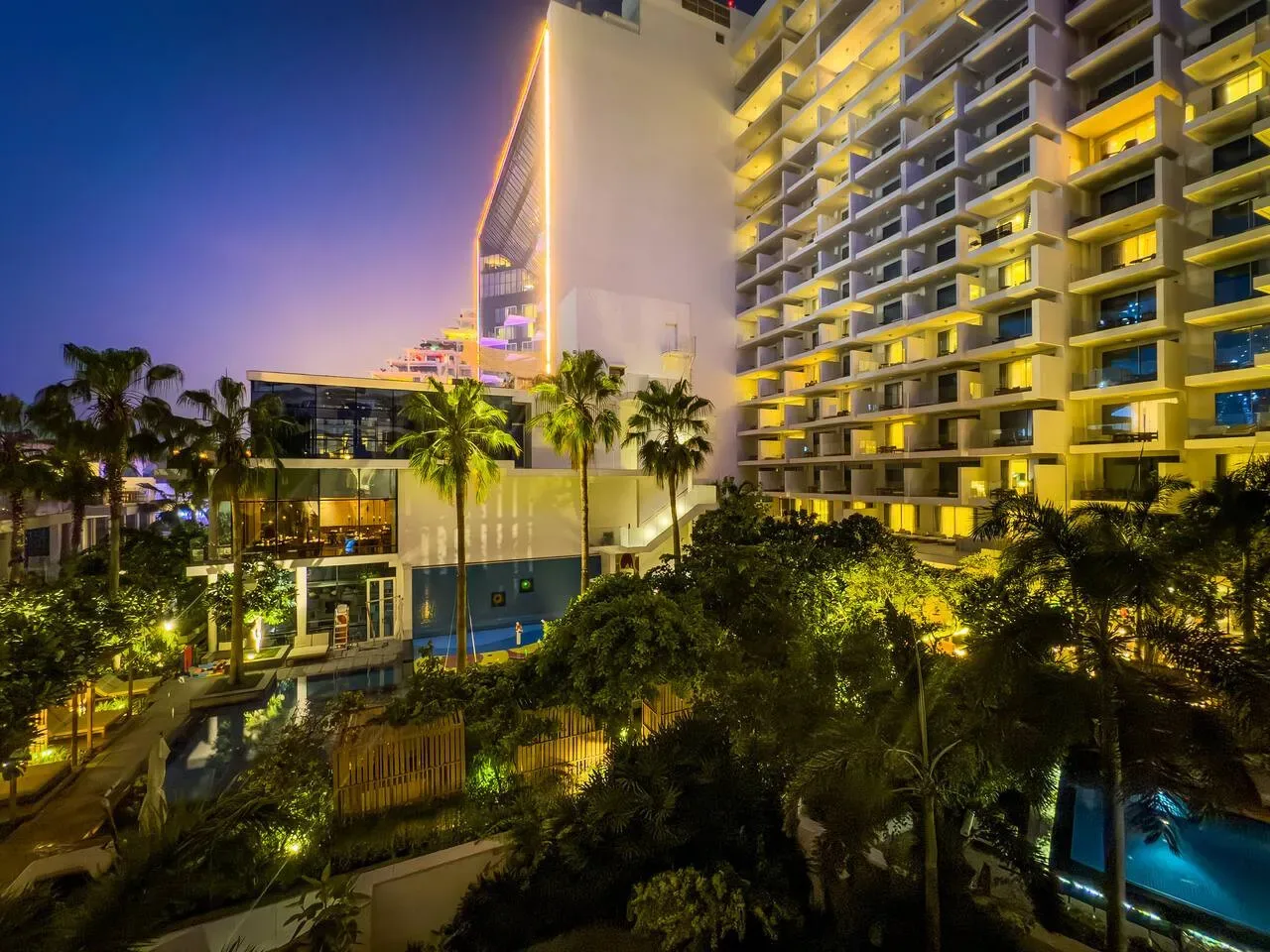 Un edificio alto y moderno se ilumina sobre jardines al anochecer.