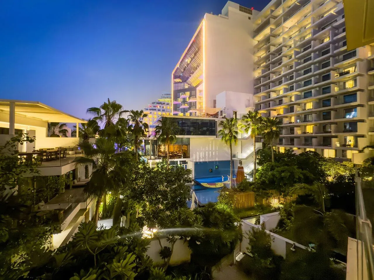 Un complejo hotelero moderno se eleva sobre un patio ajardinado con piscina al anochecer.