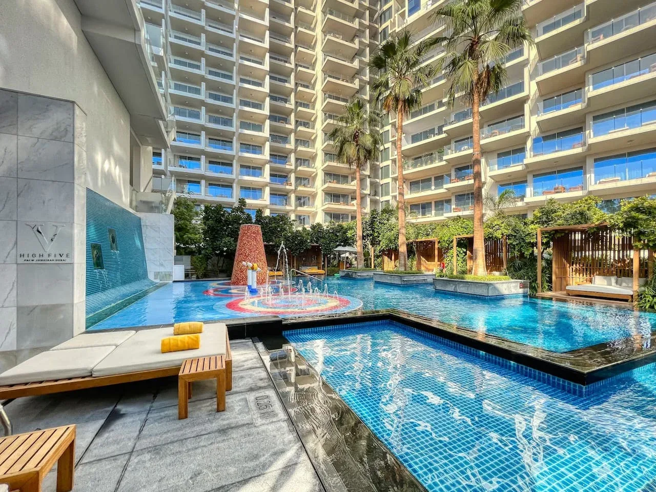 Una piscina en un patio entre edificios altos, con palmeras y asientos de descanso.