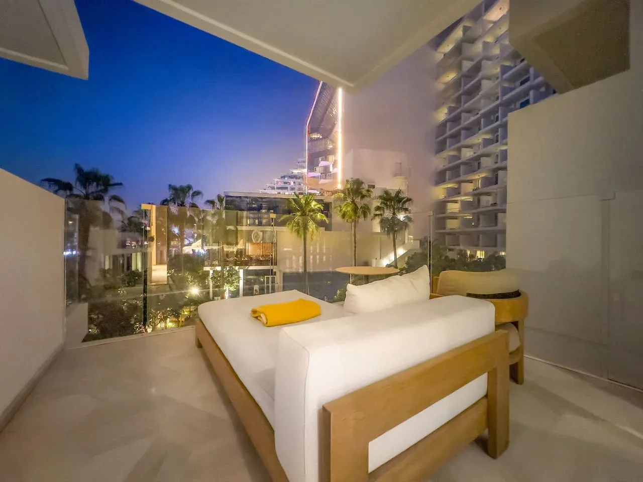 Un dormitorio con una cama blanca frente a un paisaje urbano iluminado al anochecer.