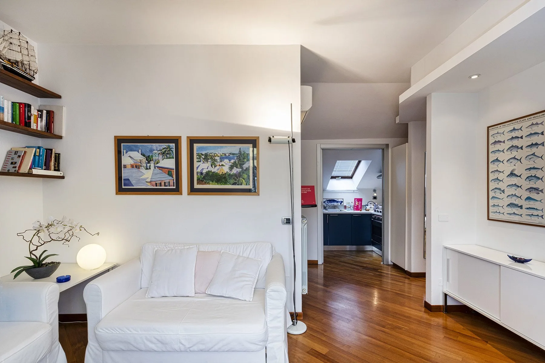 Una sala luminosa con asientos blancos y cuadros enmarcados, conectada con una cocina.