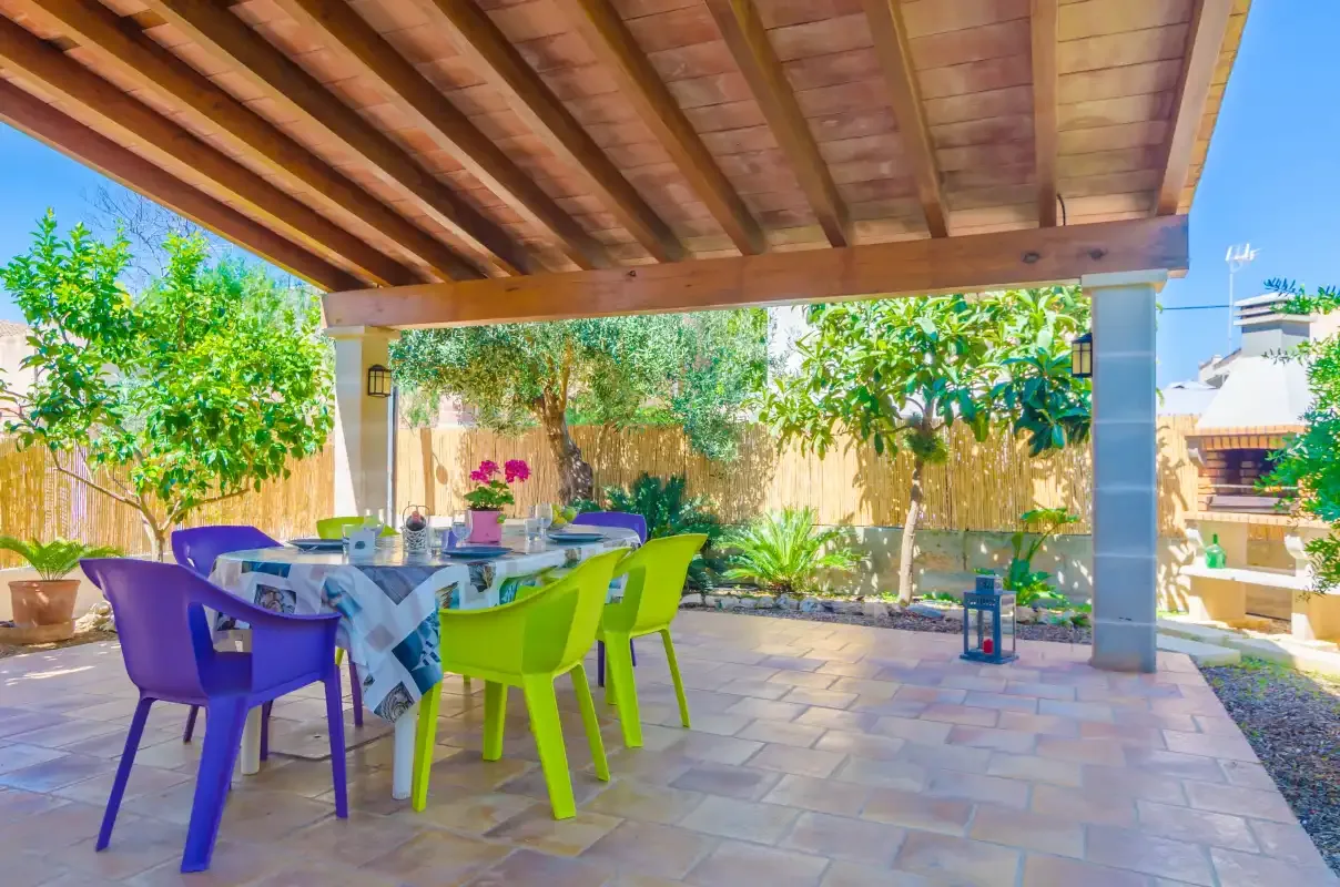 A covered patio has a dining table with colorful chairs and potted plants.