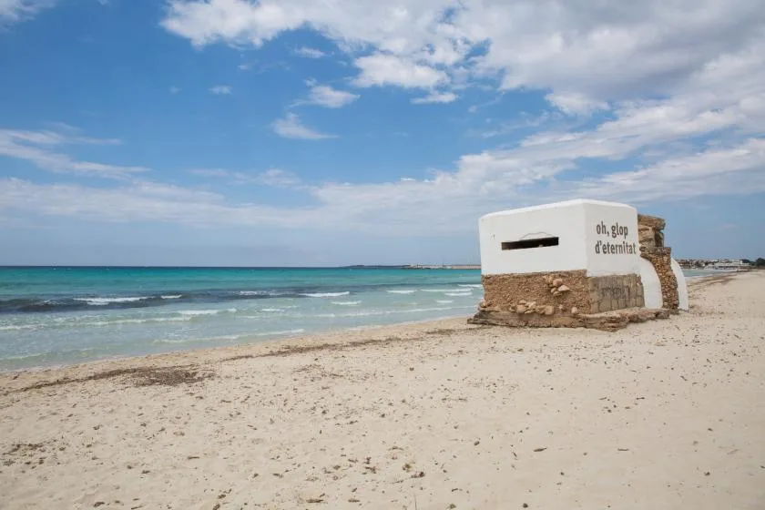 A sandy beach beside turquoise water, with a small structure near the shore.