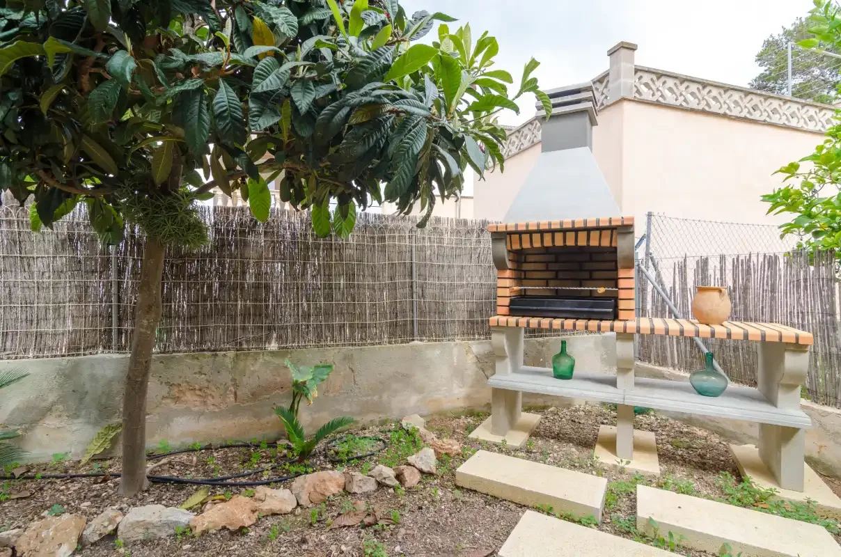 An outdoor brick barbecue stands beside a reed fence and leafy plants.