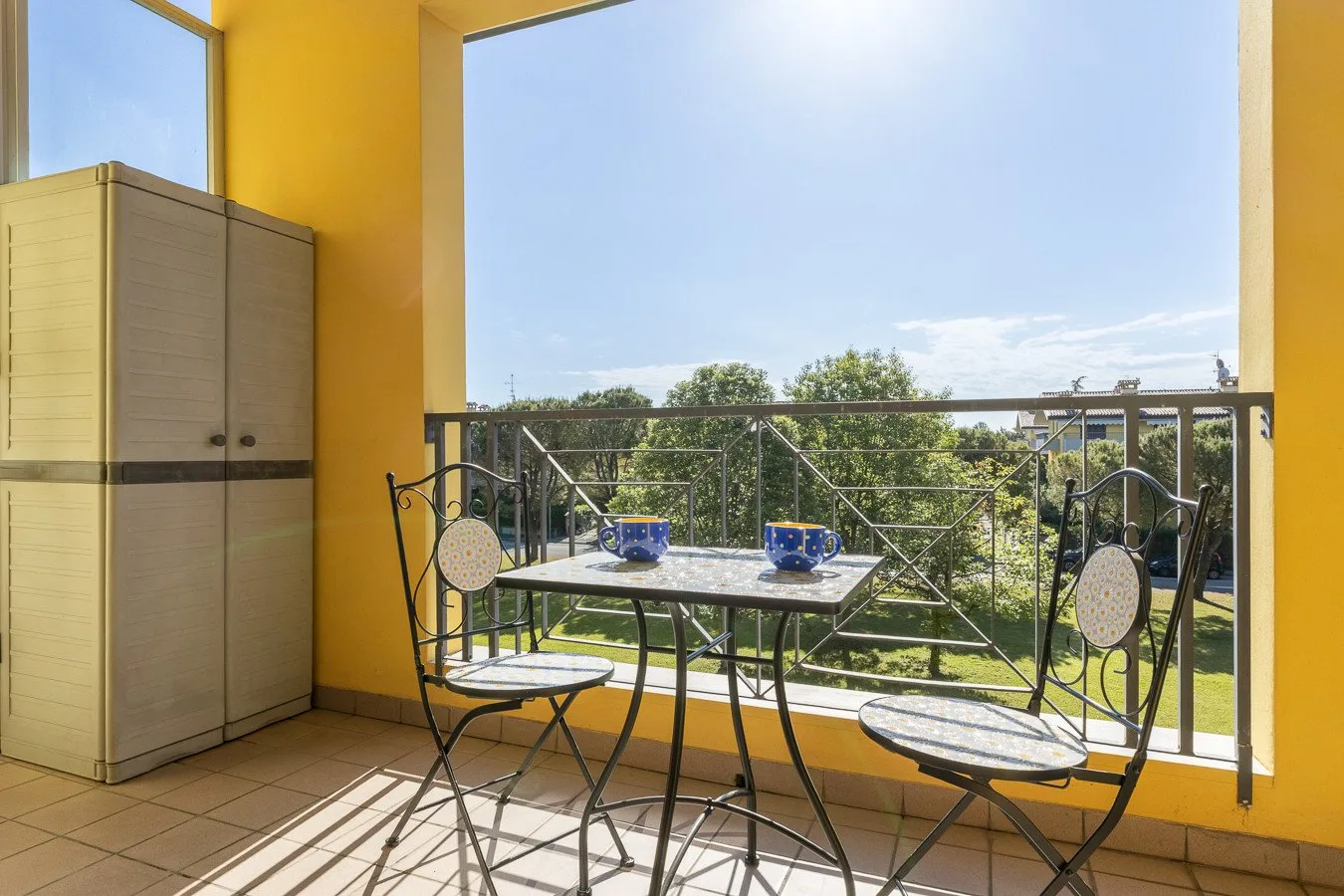 Un balcón soleado con una mesa pequeña y asientos frente a árboles verdes.