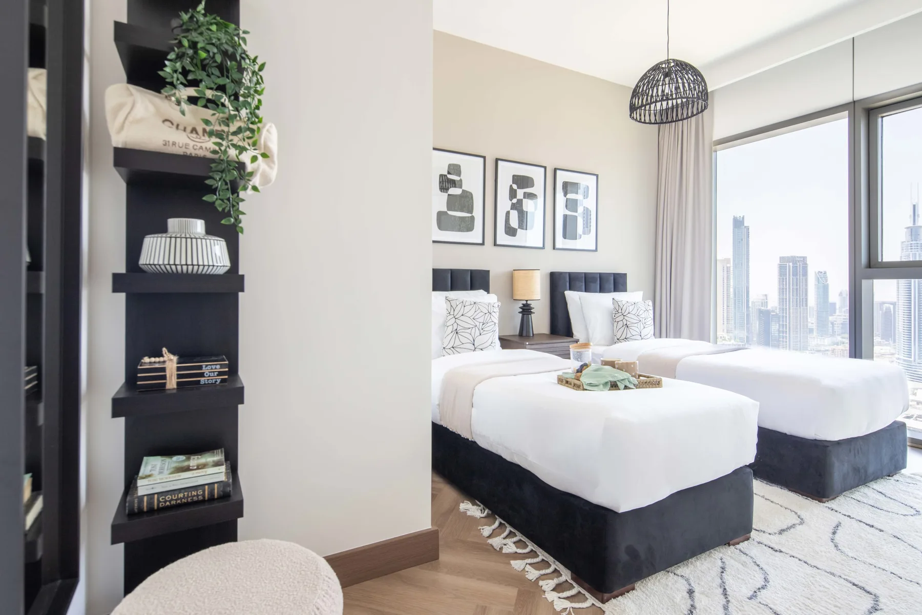 Un dormitorio luminoso con dos camas, cuadros enmarcados y grandes ventanales con vistas a la ciudad.
