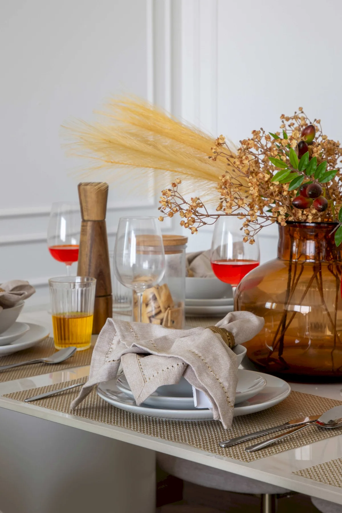 Una mesa de comedor está puesta con platos, copas y un jarrón con flores secas.