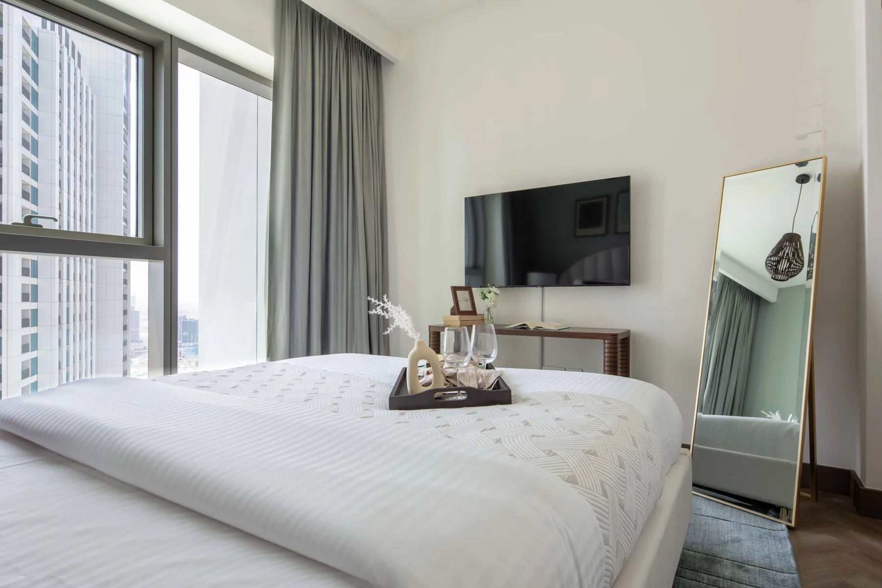 Un dormitorio luminoso con una cama bien hecha, grandes ventanas y un televisor en la pared.
