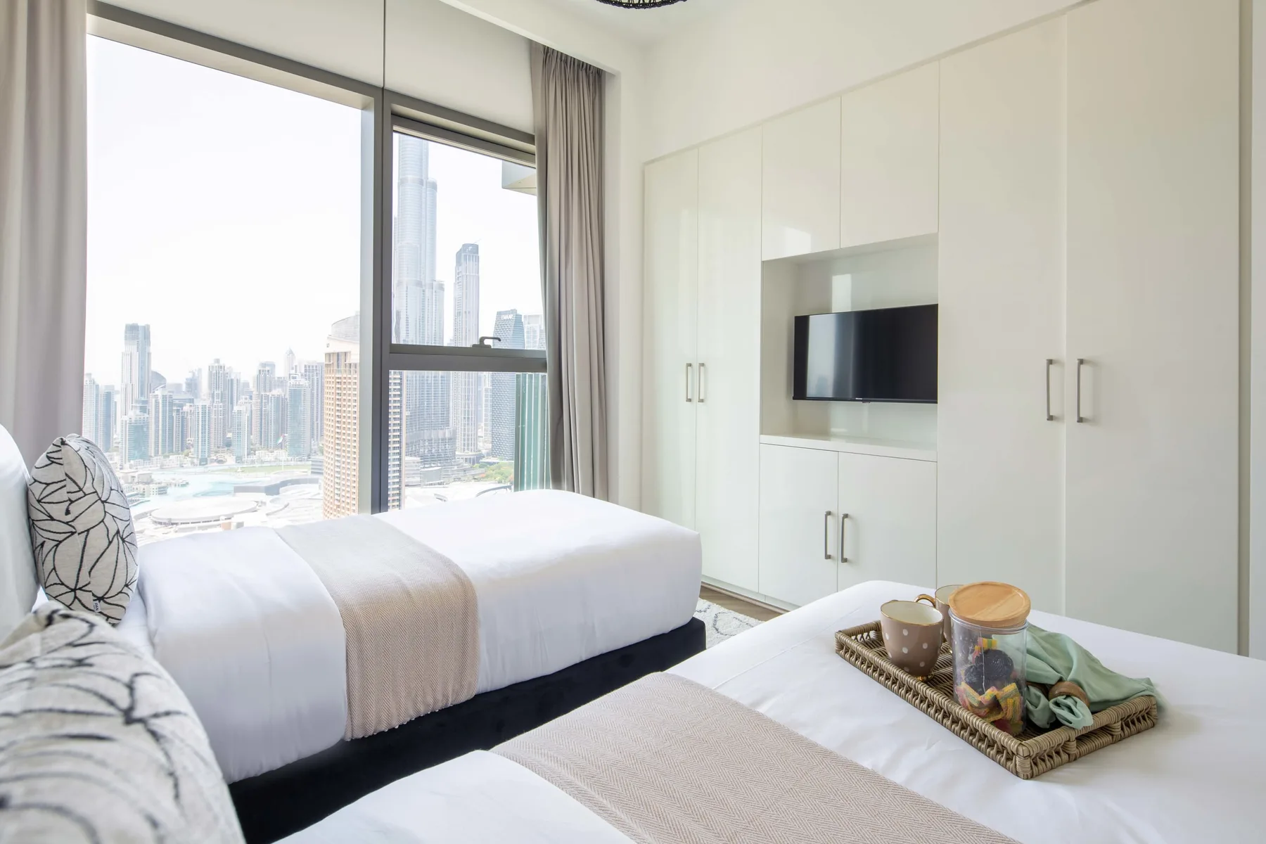 Un dormitorio luminoso con camas blancas, armarios empotrados y grandes ventanales con vistas a la ciudad.