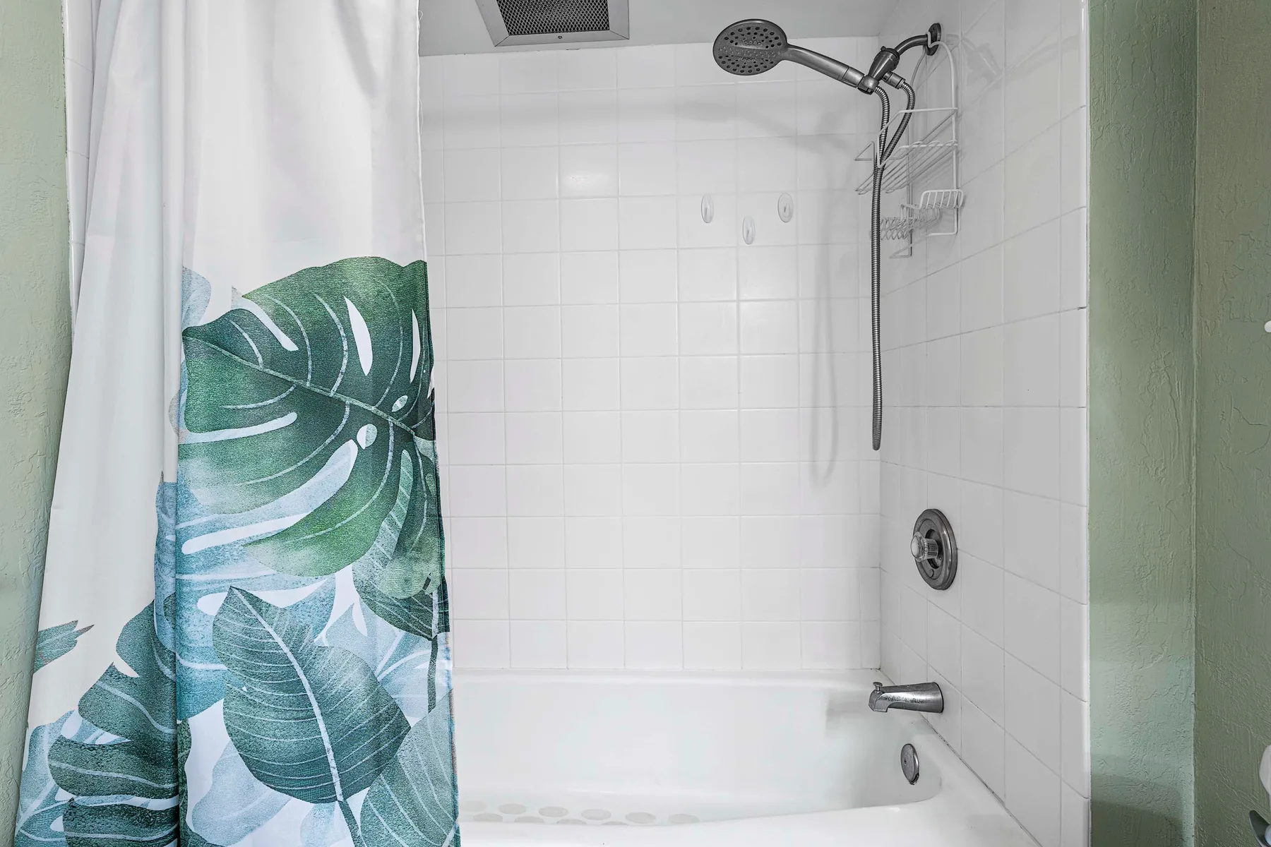 Una bañera alicatada con ducha queda detrás de una cortina con un estampado de hojas verdes.