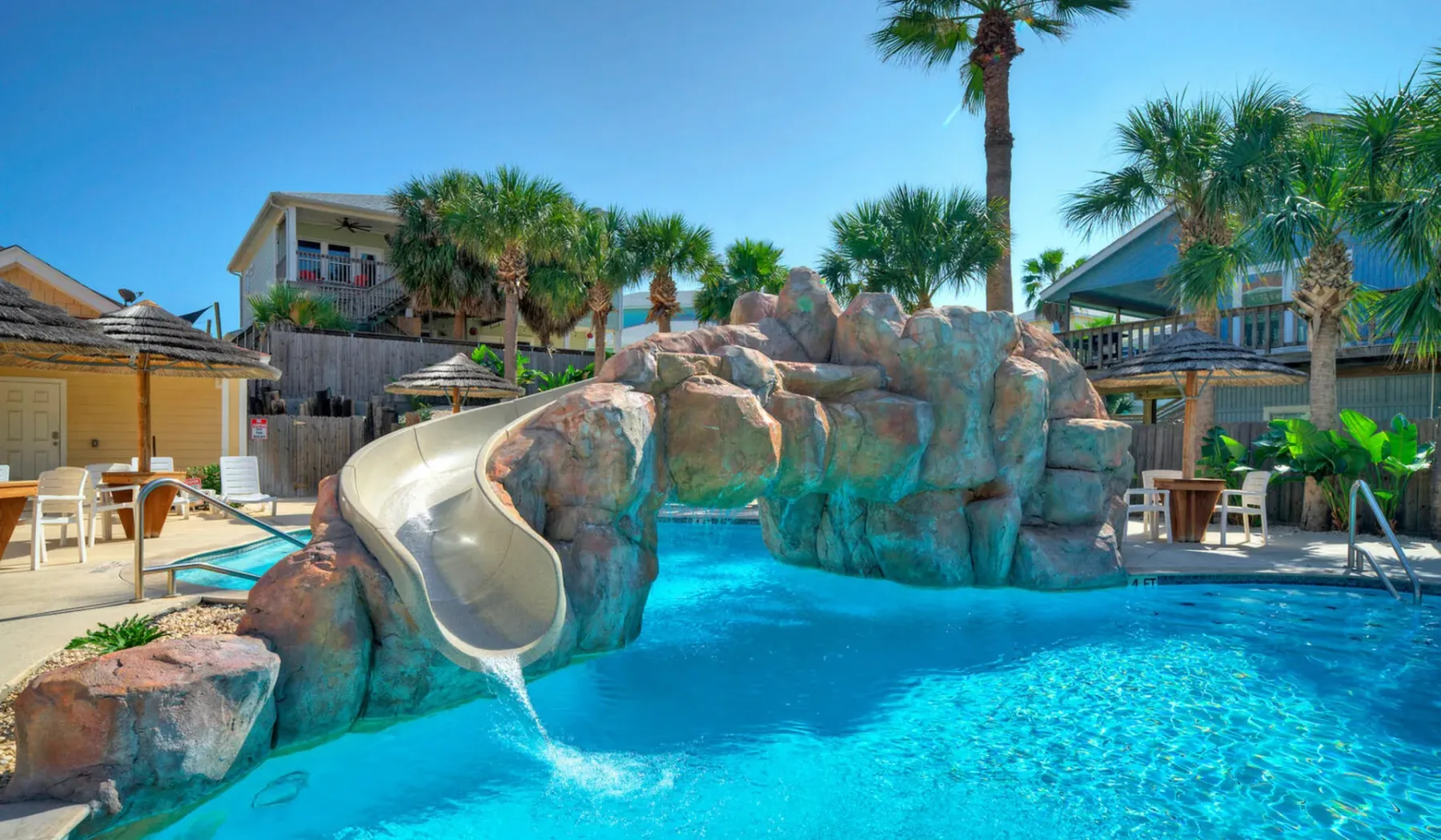 Una piscina exterior luminosa tiene una formación rocosa y un tobogán curvo.