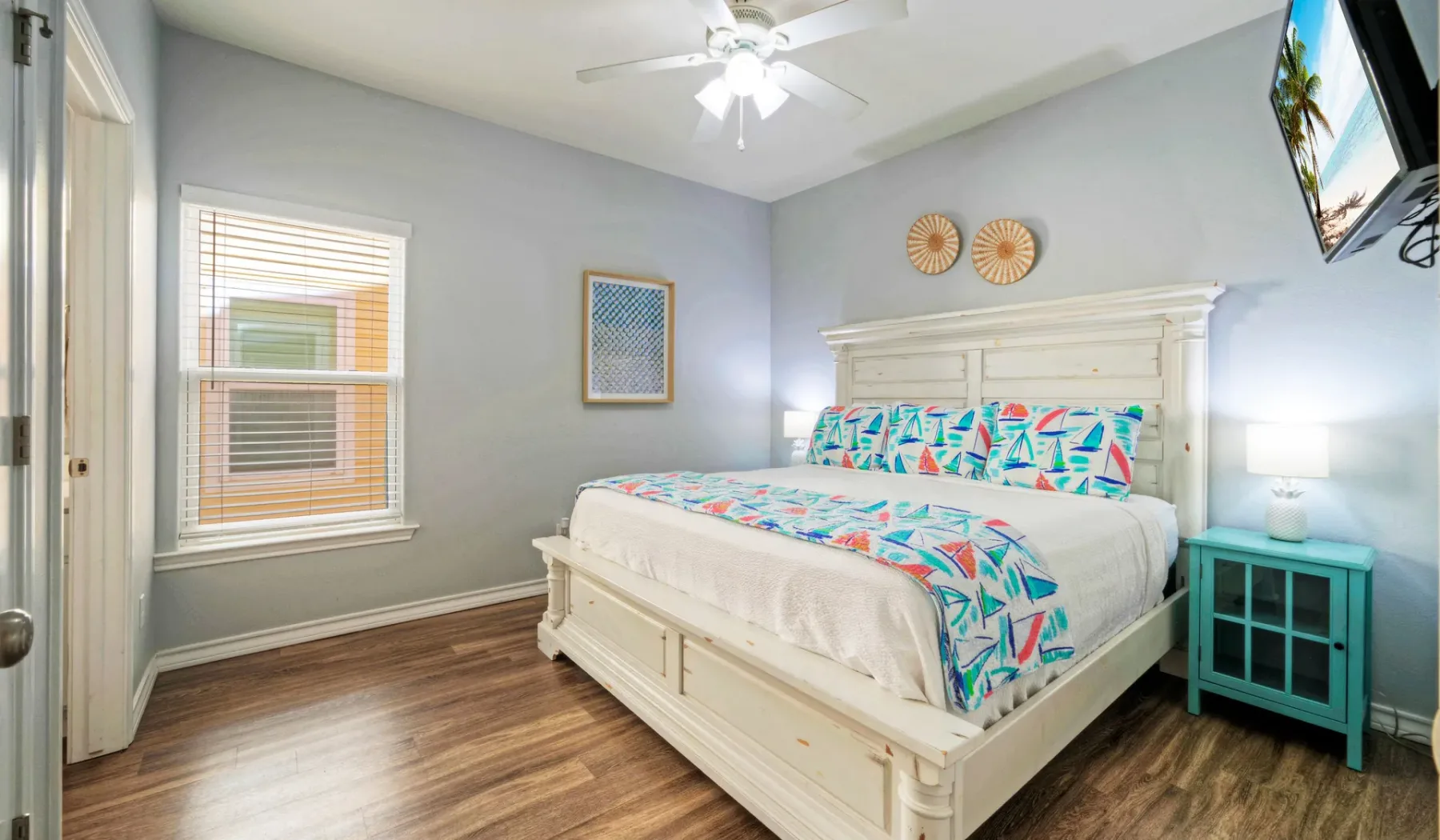 Un dormitorio con una cama blanca, ropa de cama colorida y una mesita turquesa.