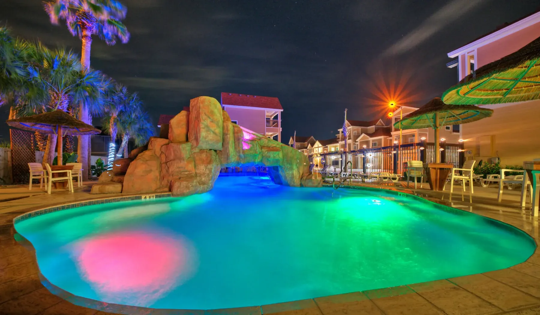 Una piscina iluminada por la noche, con formaciones rocosas y asientos cerca.