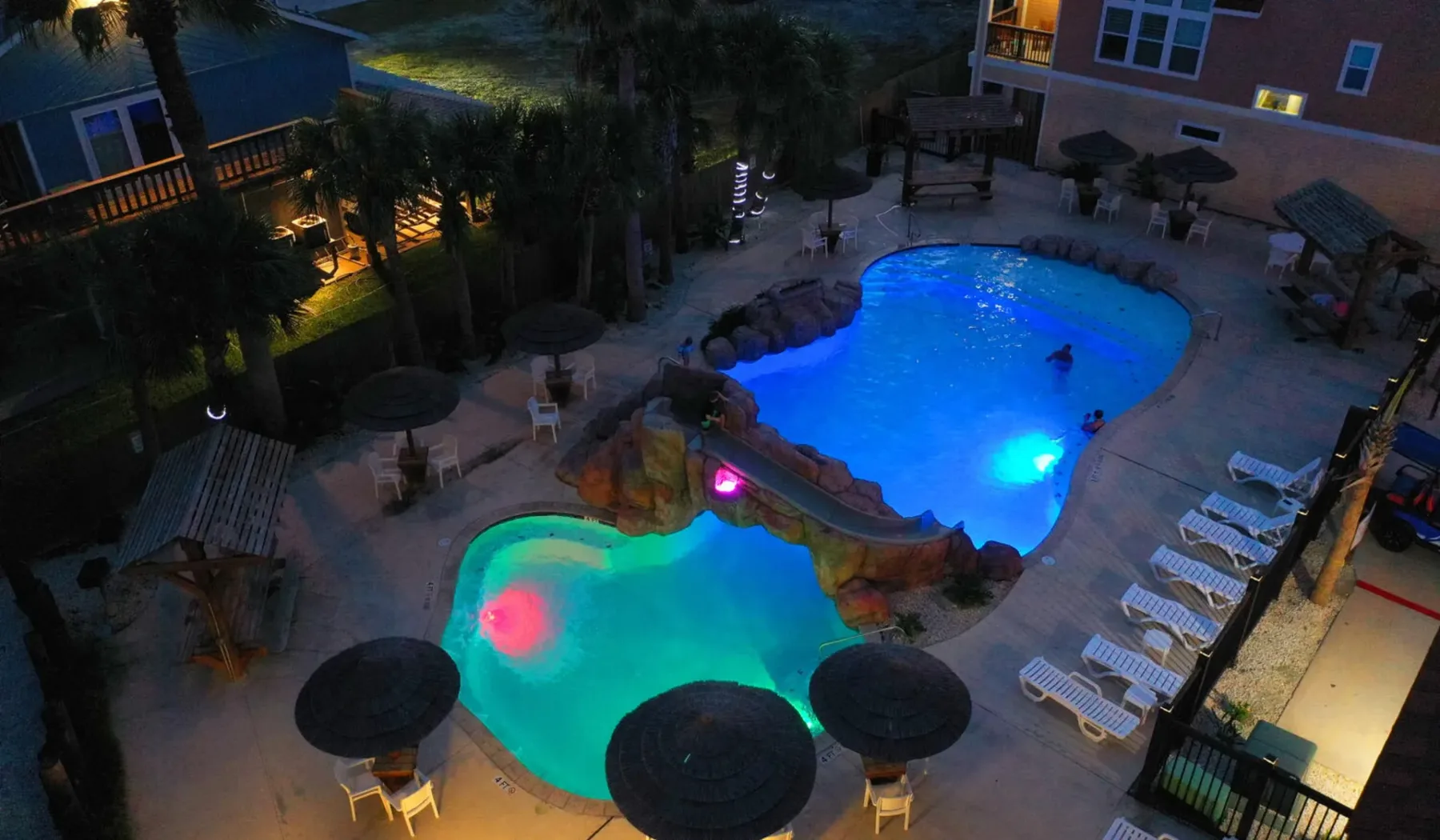 Una piscina iluminada y otra más pequeña contigua están en una zona exterior pavimentada de noche.