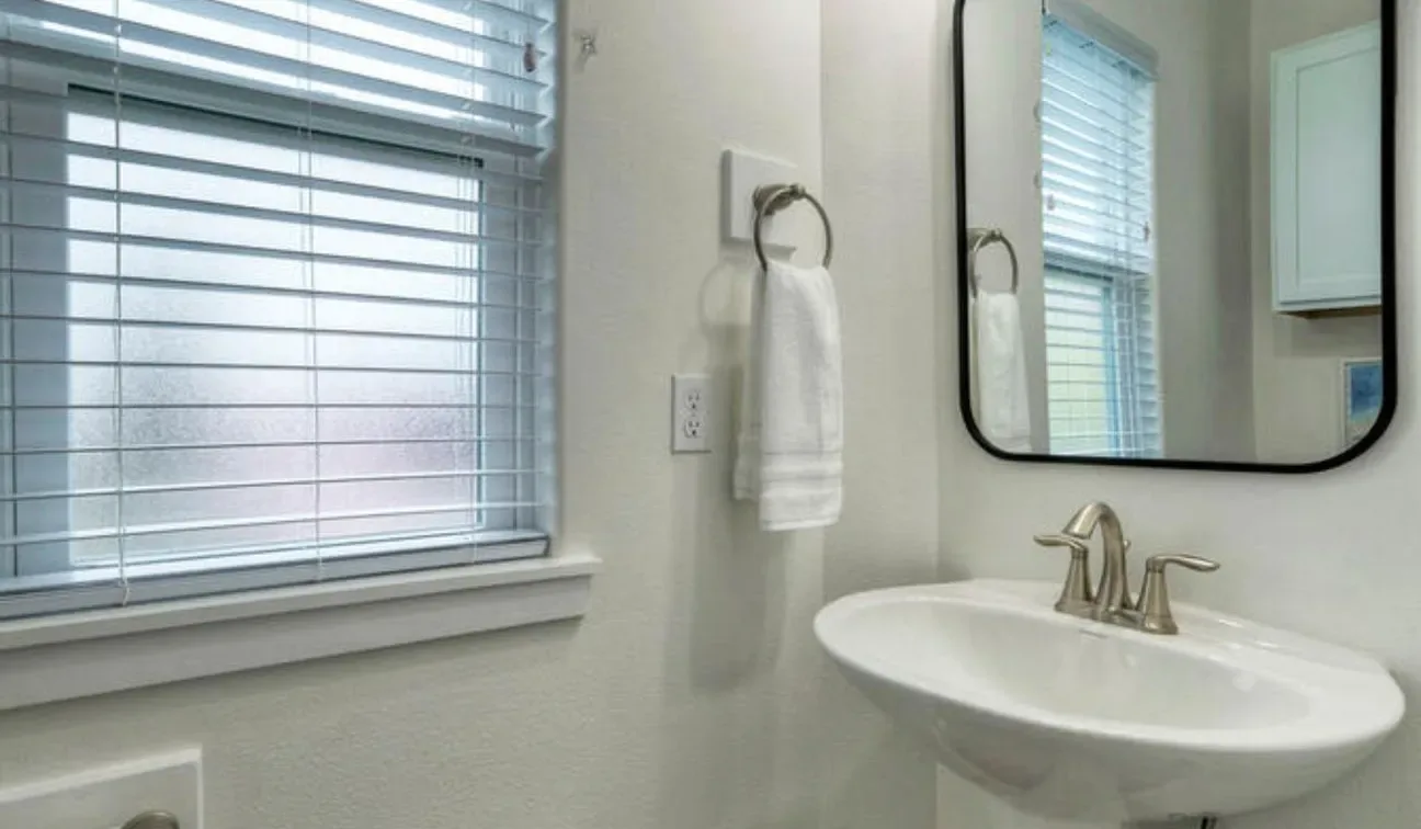 Un baño luminoso con lavabo de pared, espejo y persianas en la ventana.