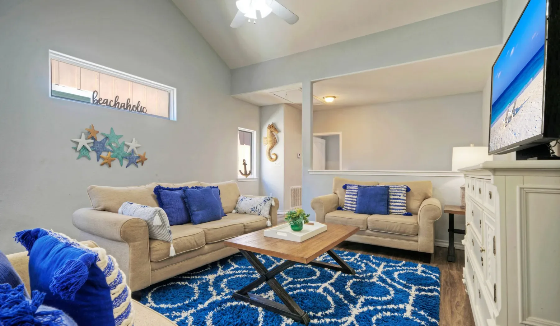 Una sala de estar con asientos beige, una alfombra azul estampada y un televisor.