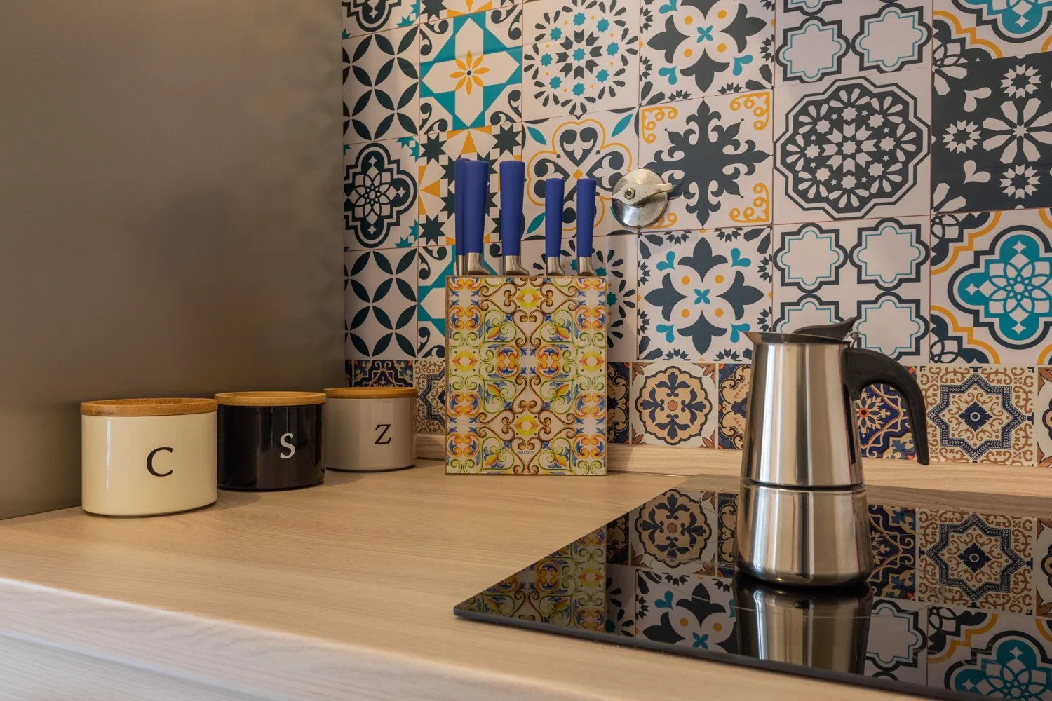 Una encimera de cocina con un bloque de cuchillos y una tetera frente a azulejos estampados.