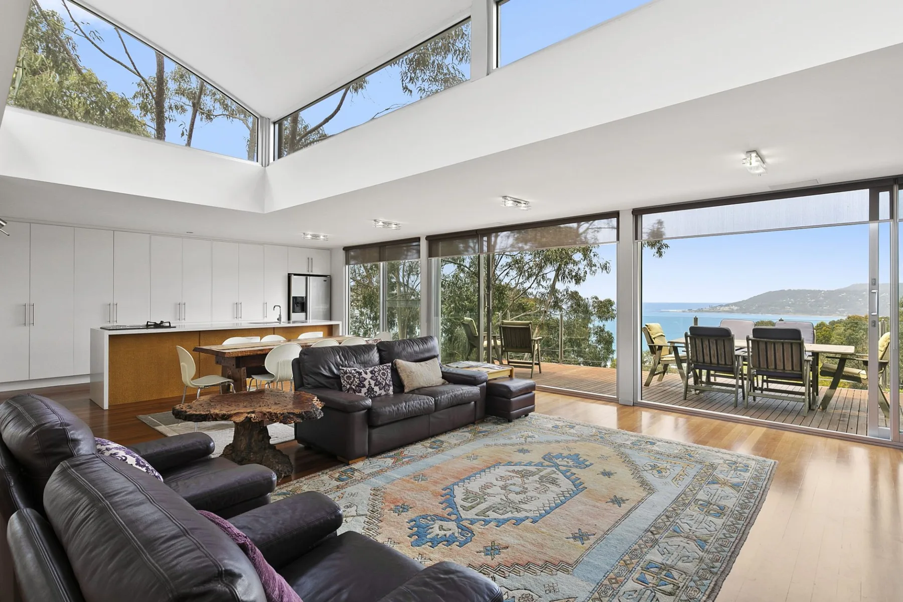 A bright open-plan living area with seating, a kitchen, and views through large windows.