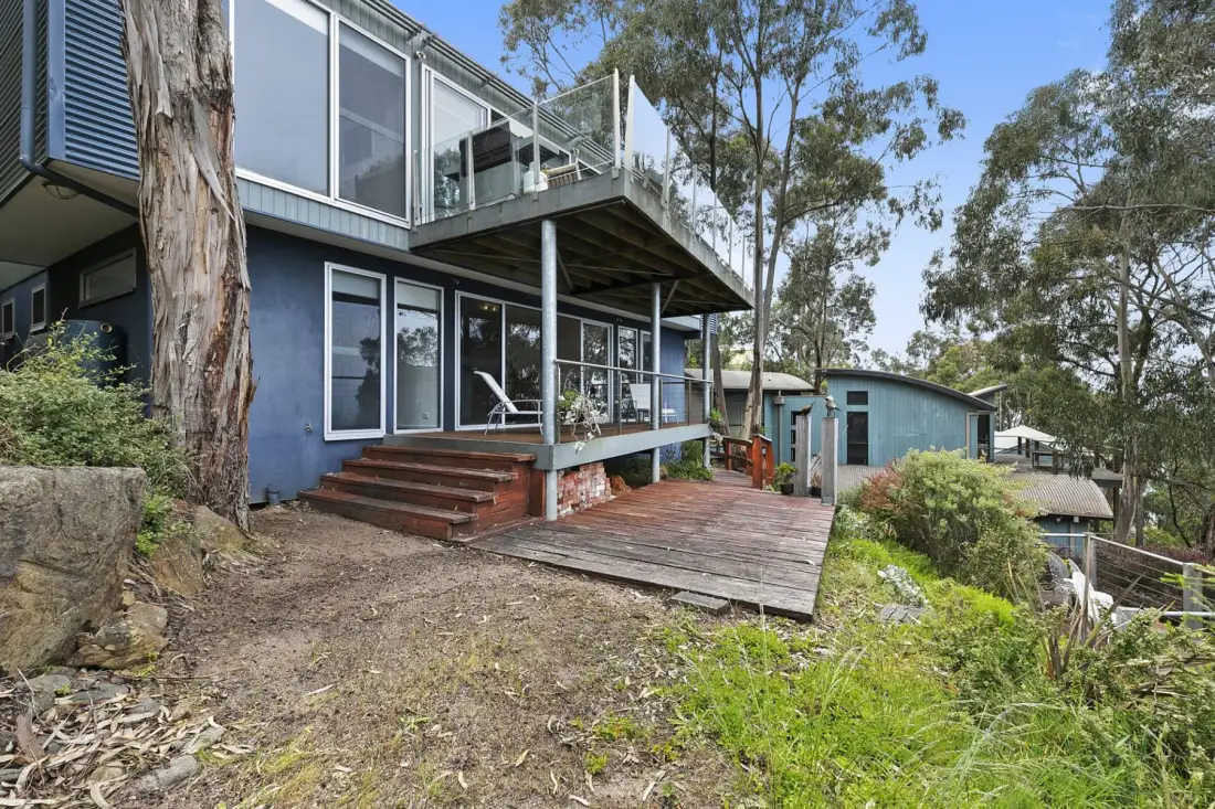 A multi-level house with a raised deck sits among trees on a sloping lot.