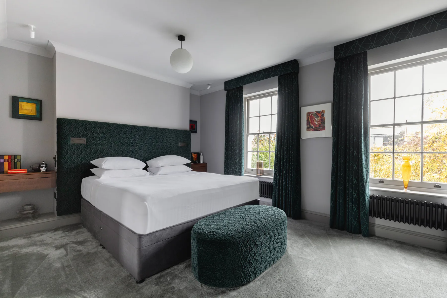 Un dormitorio con una cama bien arreglada y ventanas altas con cortinas oscuras.
