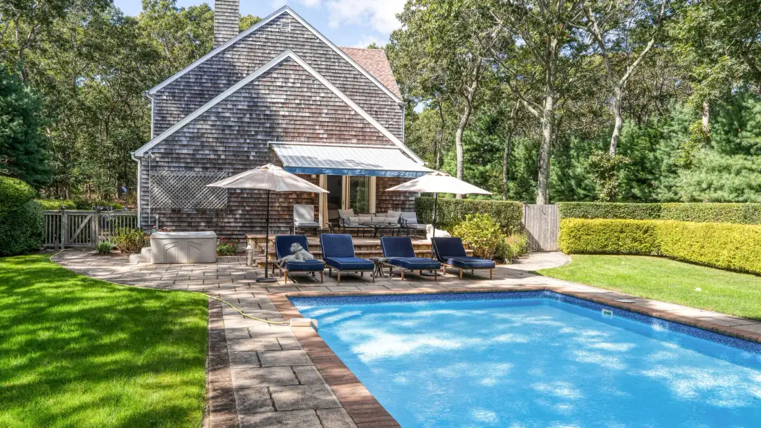 Una piscina en el patio junto a una casa, con asientos y sombrillas en la terraza.