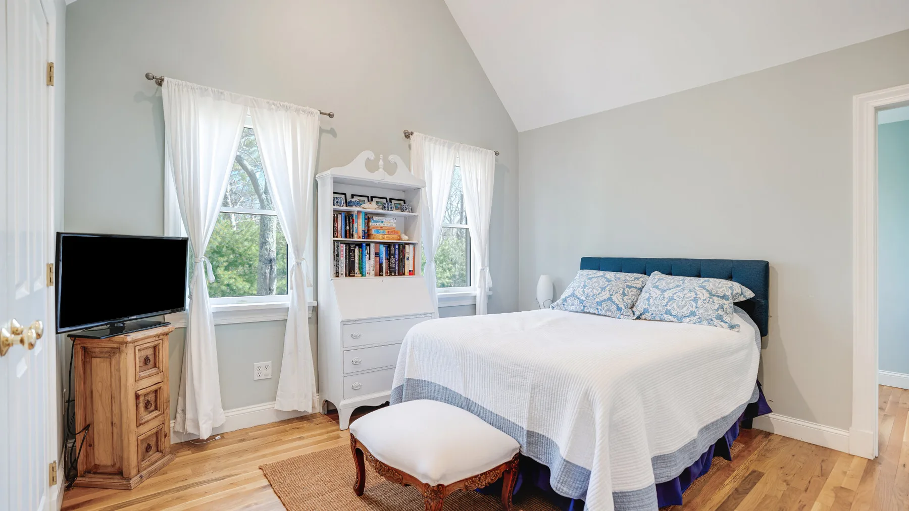 Un dormitorio luminoso con una cama, dos ventanas y una pequeña estantería.