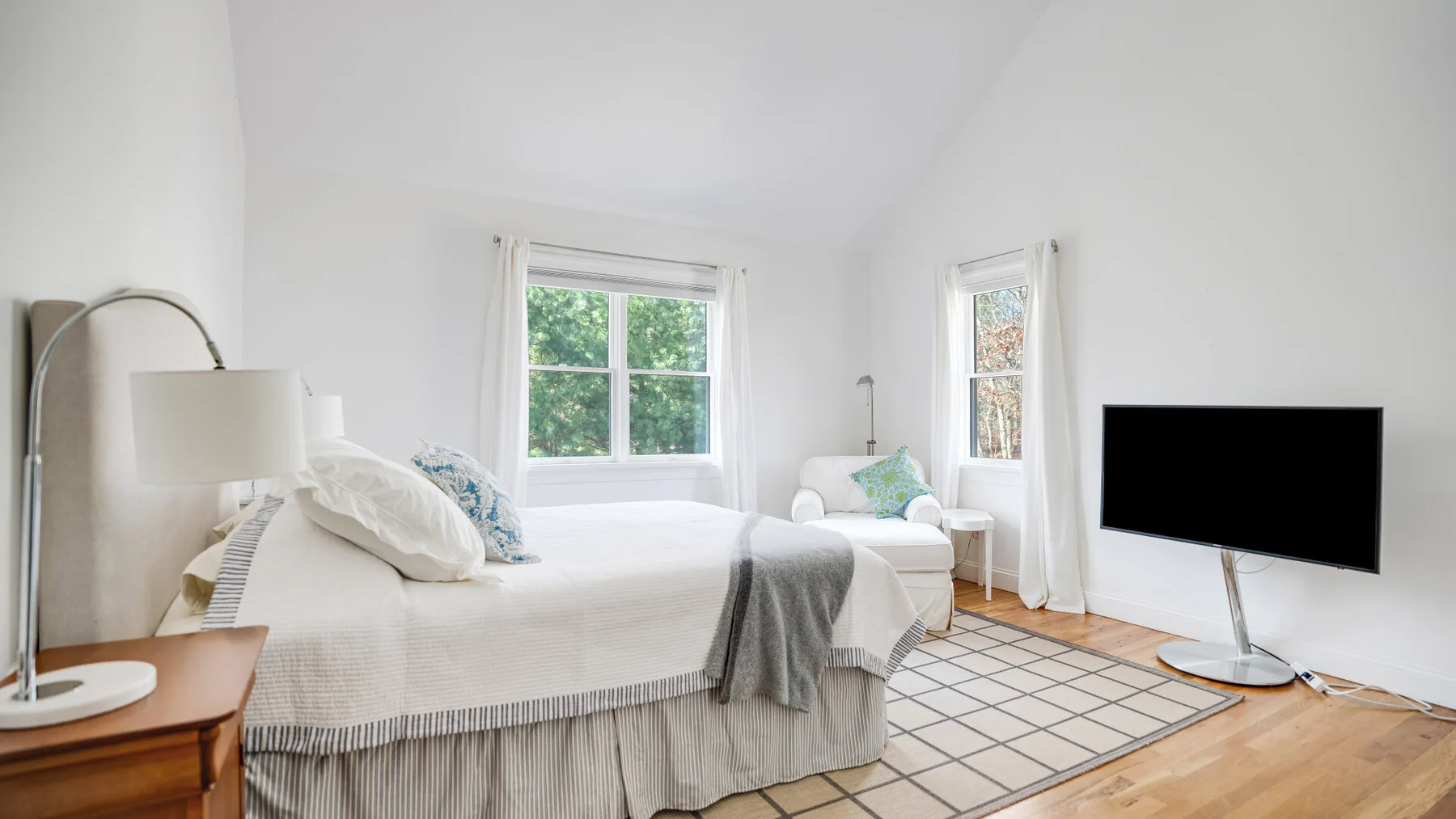 Un dormitorio luminoso con una cama, dos ventanas y un televisor.