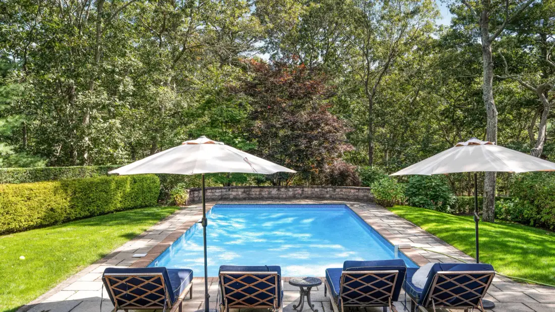 An outdoor swimming pool is surrounded by lounge seating, umbrellas, and greenery.