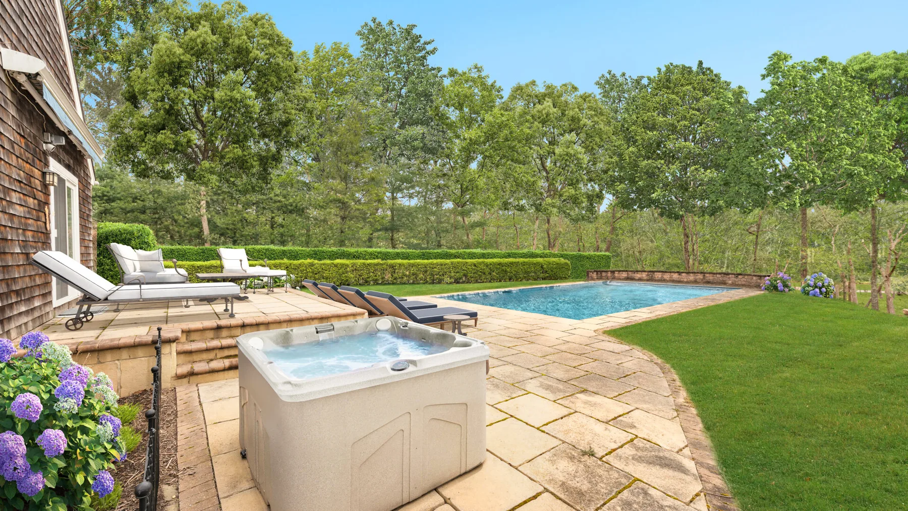 Una piscina y una bañera de hidromasaje al aire libre están junto a un patio pavimentado y un jardín.