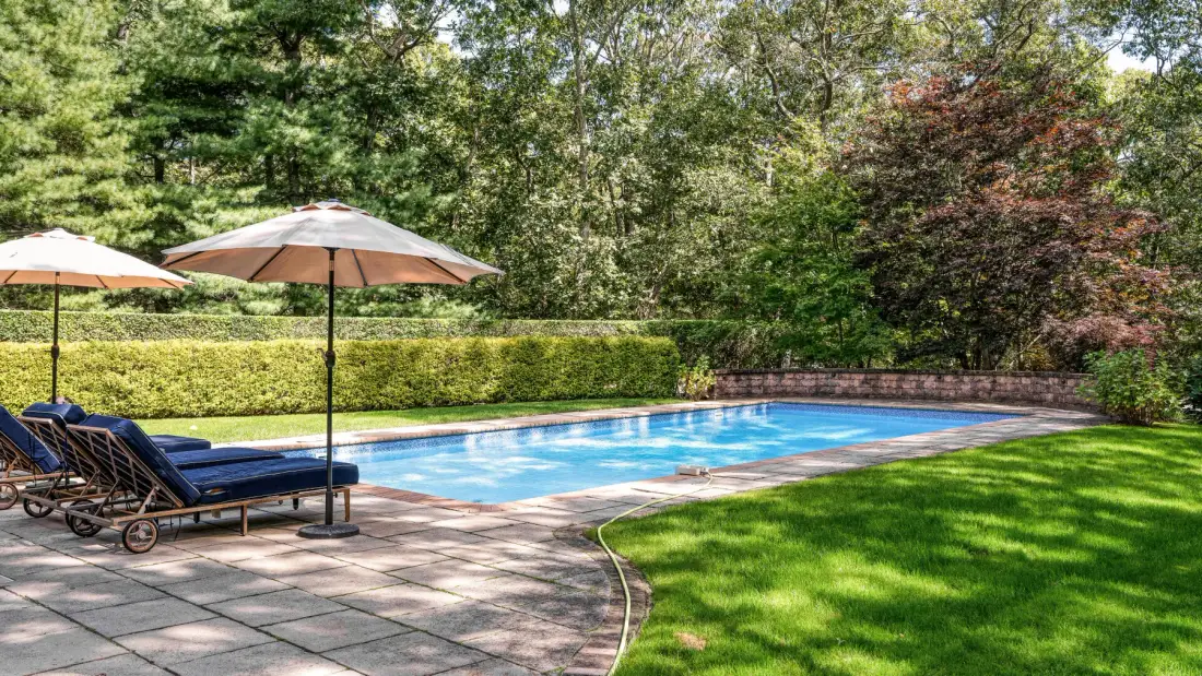 Una piscina al aire libre rodeada de un patio pavimentado, asientos y vegetación.