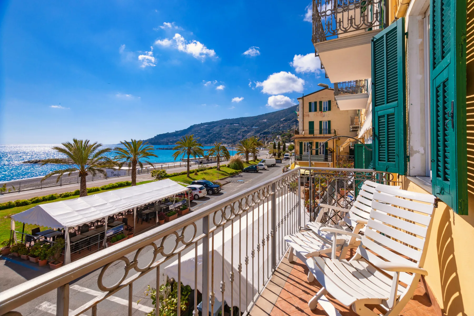 A seaside balcony overlooks a palm-lined promenade and blue water.
