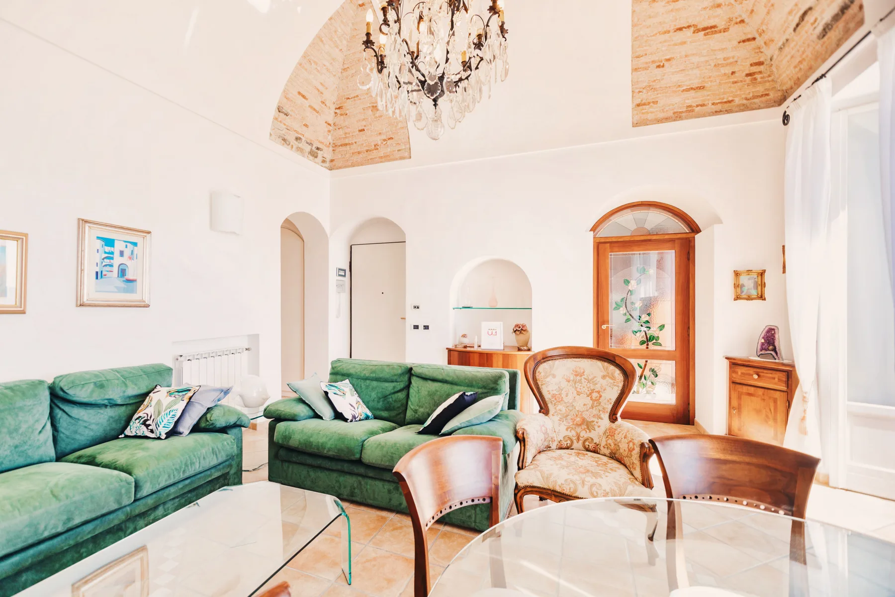 A bright living room with green seating, glass tables, and a chandelier.