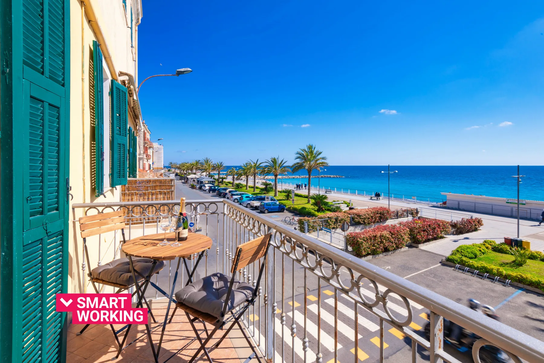 Balcony seating overlooks a palm-lined promenade and the sea.