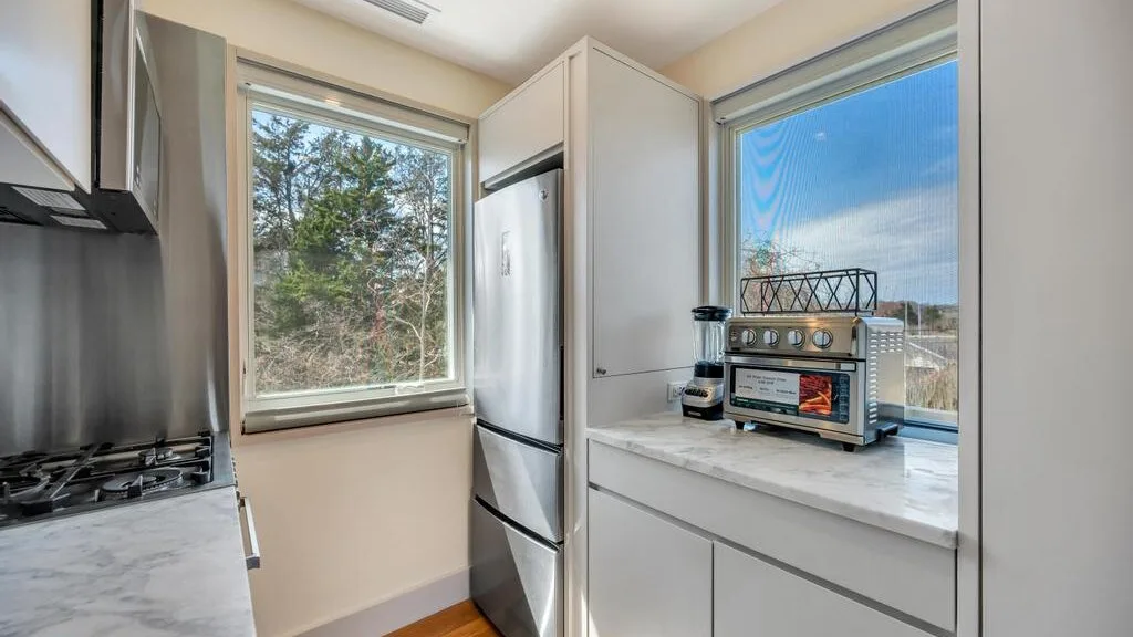 Una cocina luminosa con refrigerador, estufa y ventanas con vistas a los árboles.