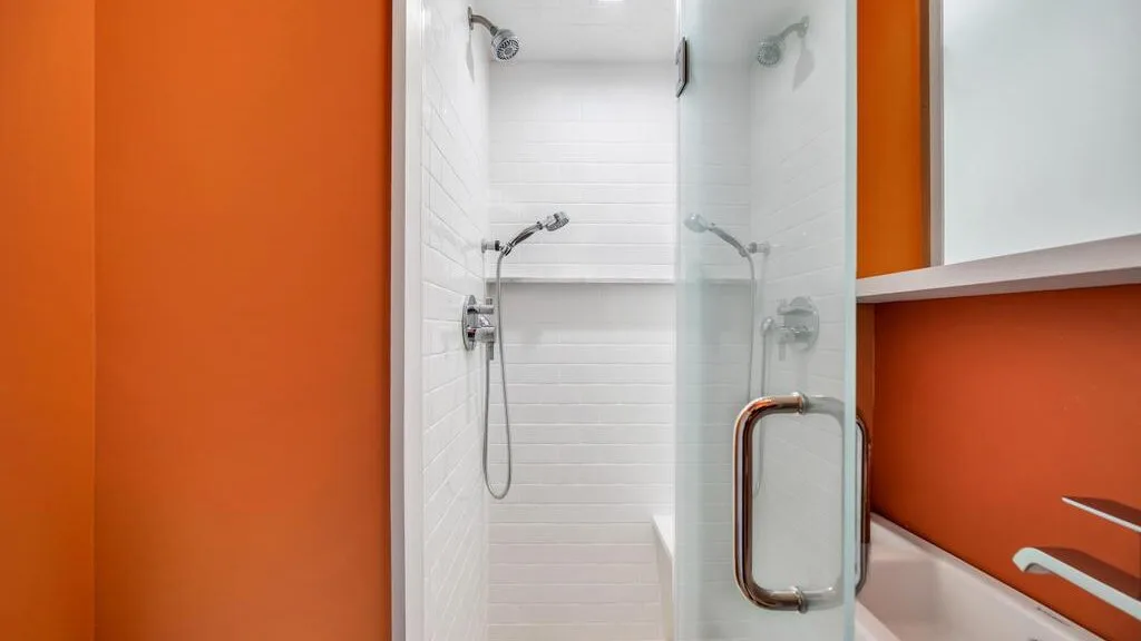 Una ducha con paredes de azulejos blancos y una puerta de vidrio junto a una pared naranja.