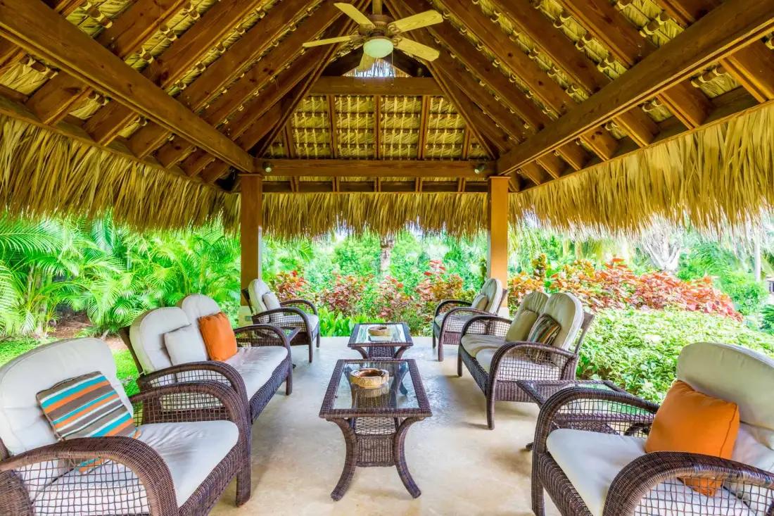 An open-sided covered seating area with cushioned chairs and low tables overlooks lush greenery.